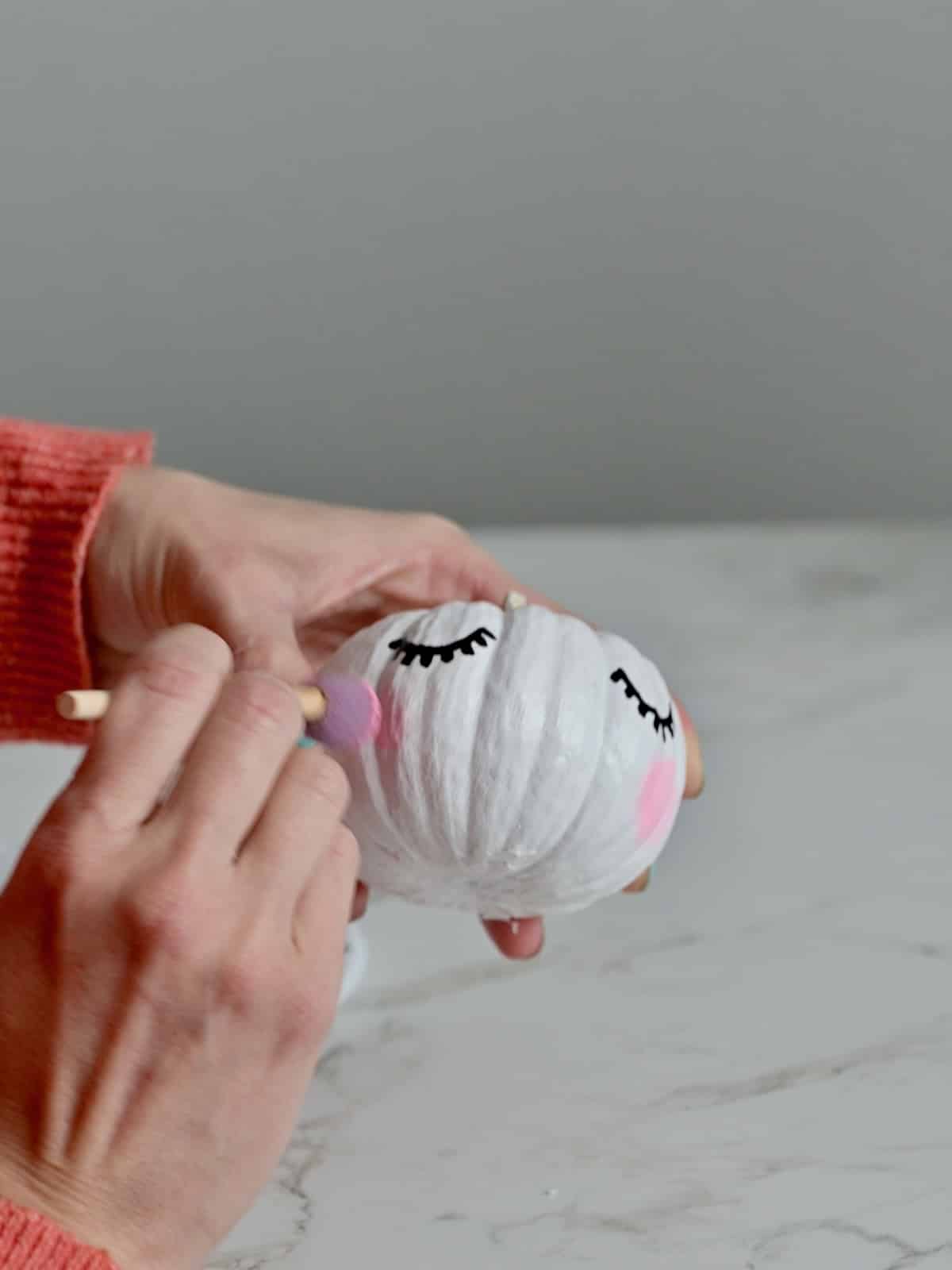 Round sponge brush adding pink paint cheeks to the pumpkin unicorn.