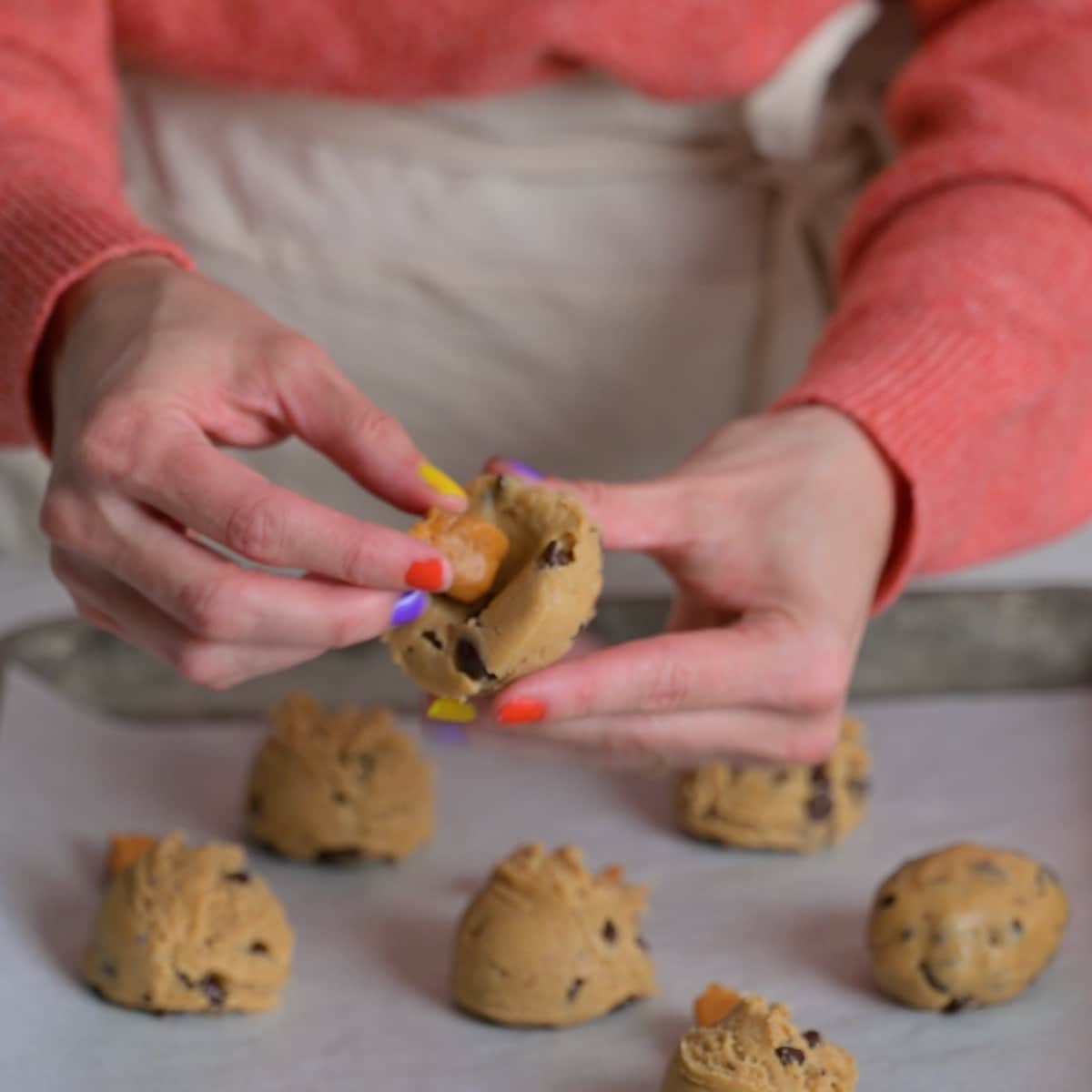 Hands pressing a caramel candy into a ball of chocolate chip cookie dough on a baking sheet.