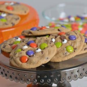 Halloween chocolate chip cookies decorated with candy eyes and colorful M&Ms on a black cake stand.