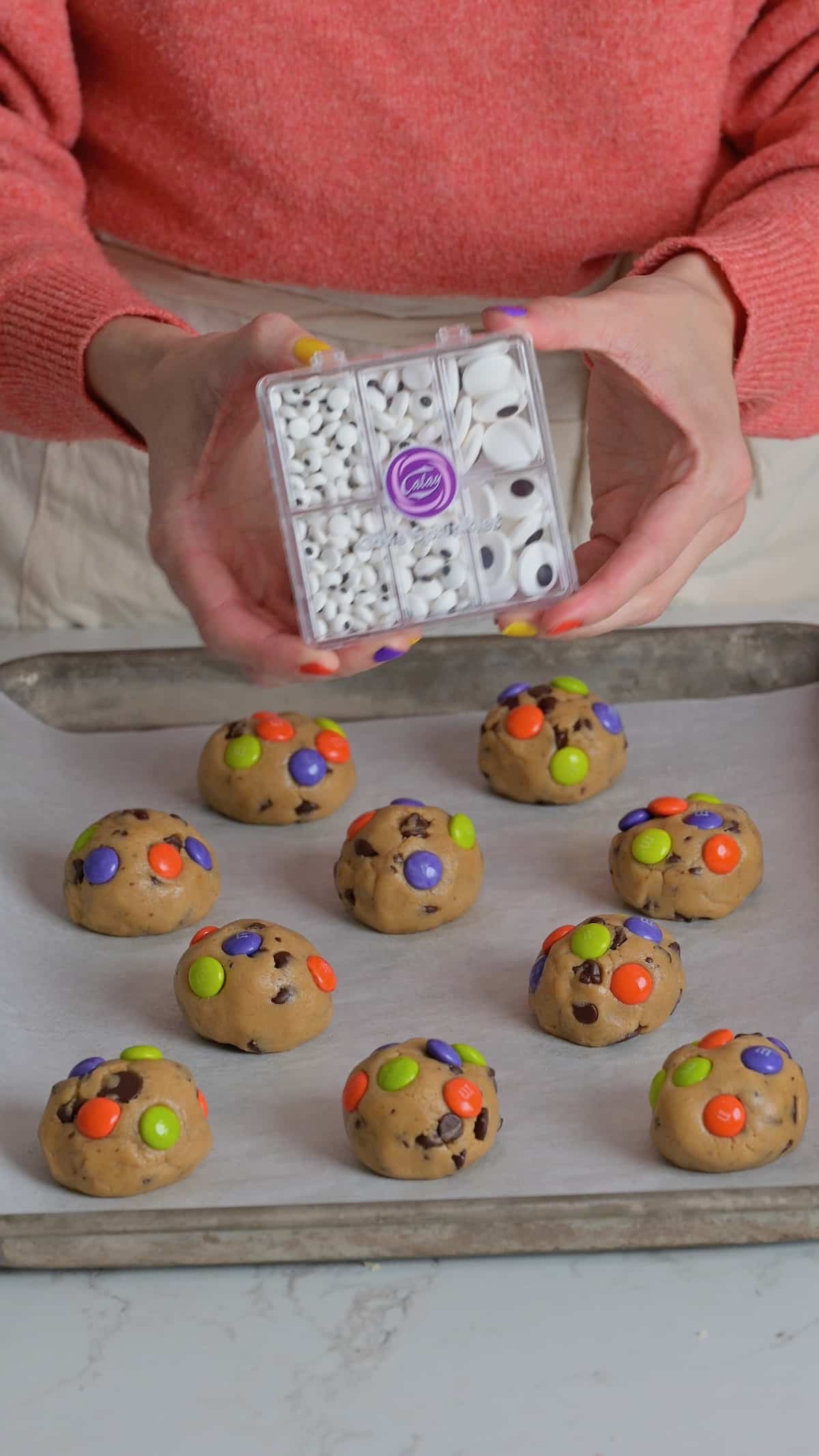 Hands holding a container of candy eyeball sprinkles above Halloween chocolate chip cookie dough balls topped with orange, green, and purple M&Ms.