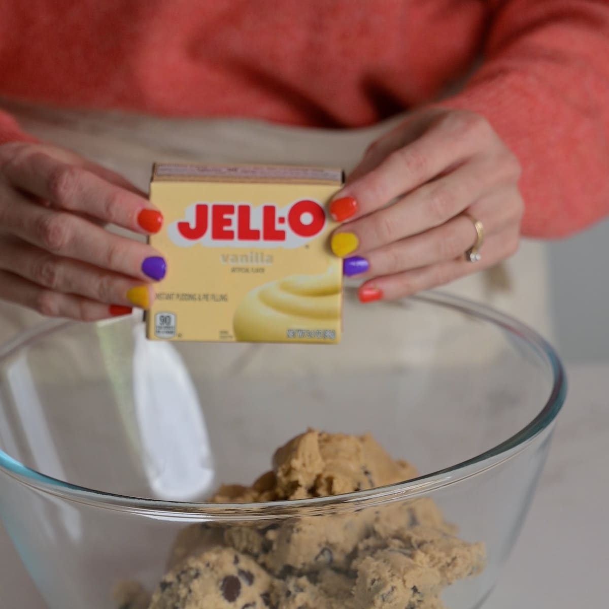 Hands holding a box of Jell-O vanilla instant pudding mix above a glass bowl of cookie dough.